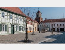 Kleiner gepflasterter Platz in einer historischen Altstadt, umgeben von Fachwerk- und Ziegelhäusern mit roten Dächern. Im Hintergrund ist ein Kirchturm zu sehen, auf dem Platz stehen junge, kahl wirkende Bäume.