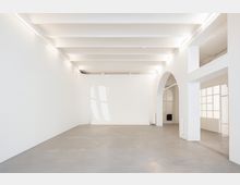 Heller, minimalistischer Innenraum mit weißen Wänden, sichtbaren Deckenbalken und glattem Betonboden. Rechts führen ein großer Rundbogen und verglaste Tür- und Fensterflächen in angrenzende Räume.