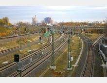 Mehrgleisige Eisenbahnstrecke mit Oberleitungen und Signalanlagen, die sich unter einer Brücke hindurchzieht; im Hintergrund sind herbstliche Bäume und moderne Gebäude einer Stadt zu sehen.