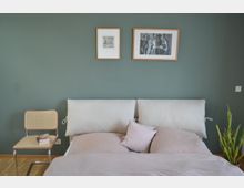 Ein Schlafzimmer mit einer hellen Bettwäsche auf einem Doppelbett mit gepolstertem Kopfteil vor einer grün gestrichenen Wand. An der Wand hängen zwei gerahmte Bilder, rechts steht eine Zimmerpflanze, links ein Stuhl mit geflochtener Sitzfläche und darauf ein Stapel Bücher.