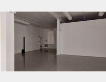 Leerer Raum mit glatten Betonboden, weißen Wänden und sichtbaren Luftrohren an der Decke. Im Hintergrund befinden sich zwei Feuerlöscher, ein Fenster mit Rollladen und eine kleine Türöffnung.