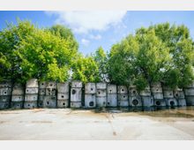 Stapel aus Betonblöcken mit runden Öffnungen vor einer Reihe dicht belaubter Bäume, auf einem betonierten, teils mit Wasser überschwemmten Platz unter einem blauen Himmel.