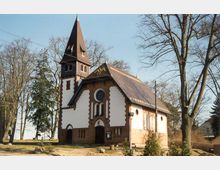 Historische Kirche mit weißer und roter Backsteinfassade, spitzem Turm und rundem Fenster über dem Haupteingang, umgeben von Bäumen in einer ruhigen Umgebung.