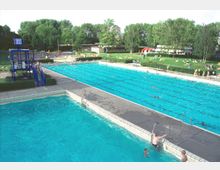 Freibad mit zwei Becken, eins zum Schwimmen und ein flacheres für Kinder, umgeben von Betonwegen und einer großen, grünen Liegewiese mit Bäumen und Menschen, die entspannen. Im Hintergrund befinden sich Umkleidegebäude und eine blaue Uhr auf einem Gerüst.