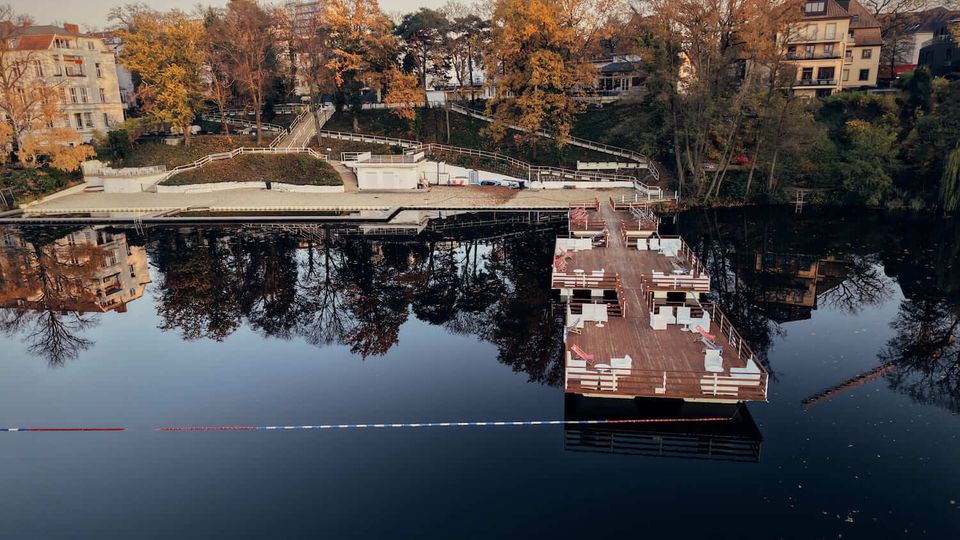 Wide view of a calm river with a large floating wooden deck and dock in the foreground, reflecting trees and nearby buildings in the water. On the opposite bank, terraced walkways and stairs run through autumn-colored trees alongside residential buildings.