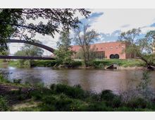 Flusslandschaft mit einer Brücke aus Holz über einem ruhigen Gewässer. Im Hintergrund ein modernes Backsteingebäude mit großen, runden Fenstern und einer Grünfläche, umgeben von Bäumen.