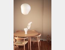 Minimal dining nook with a round wooden table and four light wood chairs on a dark wood plank floor, set against plain beige walls. A white paper pendant lamp hangs overhead, and a vase with pale flowers sits centered on the table.
