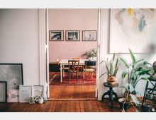 Heller Wohnraum mit Holzboden, der durch eine offene Tür einen Blick in ein Esszimmer freigibt. Dort stehen ein weißer Holztisch mit unterschiedlichen Stühlen auf einem roten Teppich; an der Wand hängen drei Schwarz-Weiß-Bilder, und Zimmerpflanzen schmücken beide Räume.