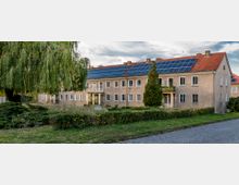 Wohngebäude mit zwei Etagen und roten Ziegeldächern, auf denen Solarpanels installiert sind. Im Vordergrund stehen eine große Weide, ein kleiner Gartenbereich und eine gepflasterte Straße.