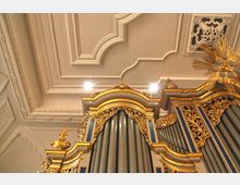 Prunkvolle Orgel mit goldenen Verzierungen und blauen Elementen vor einer reich dekorierten, weißen Stuckdecke in einem Kircheninnenraum. Im oberen Bildbereich sind ornamental gestaltete Lüftungsgitter und Beleuchtungselemente erkennbar.