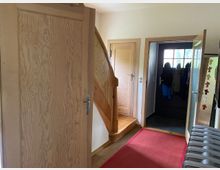 Innenansicht eines Hausflurs mit Holztüren und einer Holztreppe, die nach oben führt. Im Hintergrund befindet sich eine Glastür mit Haken an der Wand, an denen mehrere Jacken hängen, und vorne liegt ein roter Teppich auf Holzboden.