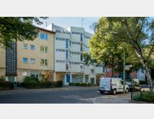 Mehrstöckige Wohnhäuser mit flachen Fassaden in einer ruhigen Wohnstraße, umgeben von Bäumen. Im Vordergrund geparkte Autos und ein Bürgersteig mit Straßenschildern.