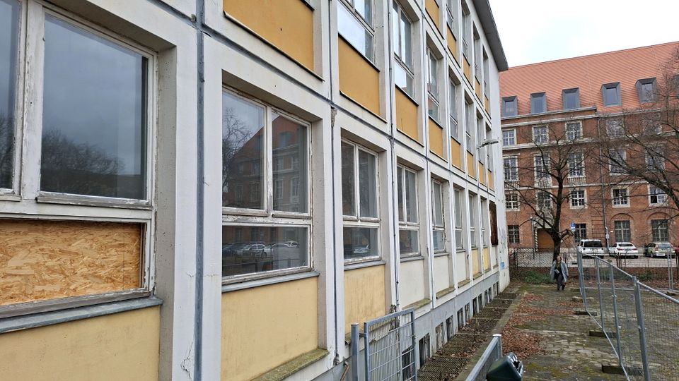 Seitenansicht eines mehrstöckigen Gebäudes mit gelb-weißer Fassade und langen Reihen rechteckiger Fenster, teils beschädigt oder mit Holzplatten abgedeckt. Daneben verläuft ein schmaler gepflasterter Weg mit Metallzäunen; im Hintergrund steht ein größeres Backsteingebäude mit rotem Dach.
