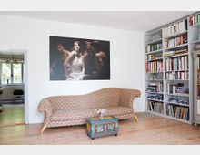 Wohnzimmer mit hellen Holzdielen, einer beigefarbenen Couch mit rotem Muster, einem kleinen bemalten Beistelltisch und einer großen Bücherwand. An der weißen Wand hängt ein großes Kunstwerk mit Porträtmotiv, und eine offene Tür führt in einen angrenzenden Raum mit Fenstern und grüner Aussicht.