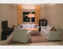 Wohnzimmer mit hellen Sofas und einem kleinen Holztisch in der Mitte. Im Hintergrund befindet sich ein Fenster mit gelben Vorhängen, daneben ein dunkler Schrank; an der linken Wand steht ein Flachbildfernseher auf einem niedrigen schwarzen TV-Schrank.