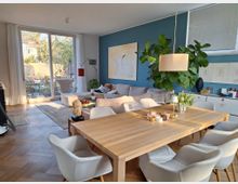 Helles Wohnzimmer mit modernem Design, bestehend aus einem großen Holztisch mit weißen Polsterstühlen im Vordergrund und einer Sitzecke mit beigem Sofa vor einer blau gestrichenen Wand. Große Fenster im Hintergrund bieten Blick auf einen Garten mit Terrasse, während Pflanzen und dekorative Elemente den Raum schmücken.