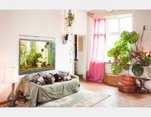 Wohnzimmer mit hellem Holzboden, einer grünen Couch mit blumigen Kissen und einem großen eingebauten Aquarium in der Wand. Ein großes Fenster mit pinkem Vorhang lässt viel Tageslicht herein, daneben stehen eine Monstera-Pflanze und gestapelte Sitzkissen.