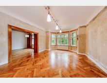 Großer, leerer Wohnraum mit Fischgrätenparkett, abgenutzten Wänden und einer großen Fensterfront mit Blick ins Grüne. Der Raum ist durch eine breite Türöffnung mit Rahmen aus Holz mit einem angrenzenden Zimmer verbunden.