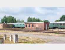 Ein kleiner, einstöckiger Backsteinbau steht neben mehreren grünen Waggons auf einem Bahngelände, umgeben von Schienen und vereinzelter Vegetation. Im Hintergrund sind Bäume und ein bewölkter Himmel zu sehen.