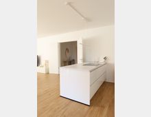 Moderne, minimalistisch gestaltete Küche mit einer freistehenden weißen Kücheninsel und integriertem Kochfeld; helles Interieur mit Holzboden, weißen Wänden und einer offenen Tür, die in einen weiteren Raum führt.
