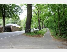 Straßenrand mit einem Fuß- und Radweg, gesäumt von hohen Bäumen und dichter Vegetation; rechts der Weg, links eine asphaltierte Straße, neben der ein Gebäude mit Ziegelfassade und Satteldach zu sehen ist.