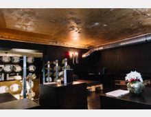 Innenraum einer Lounge mit dunklen Wänden und niedriger Decke, die teilweise abgenutzt wirkt. Es gibt Sitzmöbel auf Holzpaletten, moderne Dekoration wie Metallplatten an einer Trennwand sowie Blumenarrangements auf den Tischen, und ein sichtbarer Lüftungsschlauch an der Decke verstärkt den industriellen Stil.