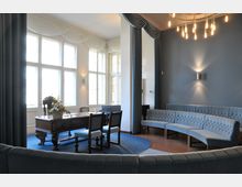 Heller Raum mit großen Fenstern, die von weißen Stoffdraperien umrahmt sind, einem dunklen Holztisch mit Stühlen im Vordergrund und geschwungenen, gepolsterten Sitzbänken an der Wand. Der Raum hat blaue Vorhänge, Wandleuchten und eine dekorative Deckenleuchte, die ein warmes Licht erzeugt.