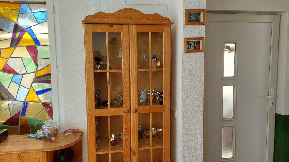 Innenbereich eines Flurs mit einem hohen Holz-Vitrinenschrank mit Glastüren, daneben eine helle Tür mit drei schmalen Milchglasfenstern. Links ist ein großes, buntes Buntglasfenster zu sehen sowie ein kleiner Beistelltisch mit Gegenständen.