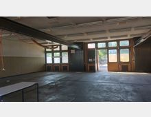 Innenraum einer leeren Werkhalle mit großen Fenstern und Glastüren, die Tageslicht hereinlassen. An der Decke befinden sich Rohrleitungen und Leuchtstofflampen, der Boden ist aus Beton, und im Hintergrund sind Heizkörper sowie ein Ausgang zum Außenbereich sichtbar.