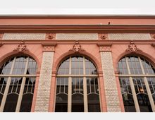 Fassade eines historischen Gebäudes mit drei großen Rundbogenfenstern, eingerahmt von rosafarbenen Stuckrahmen und weißen Ziegelpfeilern. Über den Fenstern befinden sich ornamentale Wappenreliefs und ein breites Gesims unter dem Dachrand.