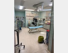 Ein heller medizinischer Behandlungsraum mit mintgrünen Wandfliesen, einem Untersuchungstisch in der Mitte und mehreren medizinischen Geräten, darunter ein Deckenarm mit Monitor. Im Hintergrund befinden sich Schränke, Spender für Verbrauchsmaterialien und Waschbecken.