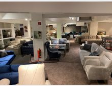 Großer Möbelgeschäft-Innenraum mit verschiedenen Sofas und Sesseln in unterschiedlichen Farben und Stilen, die auf einem braunen Teppich präsentiert werden. Im Hintergrund sind Beleuchtung, Dekoelemente sowie Regale mit weiteren Möbelstücken zu sehen.