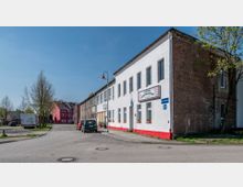 Straßenszene mit einem weißen, zweistöckigen Gebäude aus Backstein und Putz, das als Wurstproduktionsbetrieb gekennzeichnet ist, daneben weitere Gebäudereihen. Rechts und links der Straße stehen vereinzelt Bäume, im Hintergrund ein rot gestrichenes Haus.