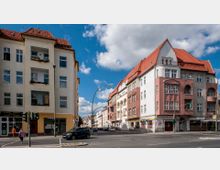 Straßenszene mit zwei angrenzenden Wohnhäusern im Altbaustil, beide mit roten Ziegeldächern und weißen Fassaden, eines davon zusätzlich mit rosa Elementen. Im Vordergrund eine Straßenkreuzung, Verkehrsampeln und geparkte Autos, bei klarem Himmel mit einigen Wolken.