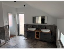 Modern attic bathroom with a sloped ceiling, gray tile flooring and wall panels, and a double vanity with two round vessel sinks beneath a wide mirror. A frosted glass door and a tall window bring in light, with a low skylight along one side wall.