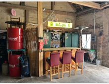 Rustikales Innenraumfoto einer umgebauten Werkstatt mit einer kleinen Bar. Im Vordergrund stehen Holzbarhocker mit roten Polstern vor einer Theke, im Hintergrund sichtbar sind alte Stein- und Holzwände, eine grüne Metallwerkzeugschrank, Dekorationen und ein leuchtendes „Bitburger Pils“-Schild.