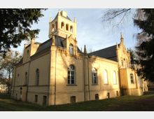 Gelb gestrichenes, historisches Gebäude mit neogotischen Architekturmerkmalen, darunter Spitzbogenfenster, Zierelemente und ein kleiner Turm. Das Gebäude steht in einer von Bäumen umgebenen, sonnenbeleuchteten Grünfläche.