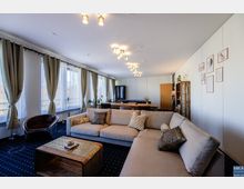 Wohnbereich mit modernem Interieur, bestehend aus einem großen beigefarbenen Ecksofa, einem Holztisch, dekorativen Kissen, und bodenlangen Vorhängen vor breiten Fenstern. Im Hintergrund befinden sich ein Esstisch, ein Regal und eine minimalistische Wanddekoration.