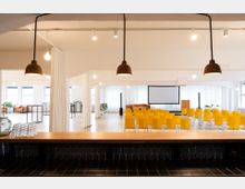 Großer, moderner Veranstaltungsraum mit gelben Stuhlreihen vor einer Bühne mit Leinwand. Im Vordergrund befindet sich eine Theke aus Holz, im Hintergrund helle Wände, große Fenster und minimalistische Einrichtung mit Pflanzen.