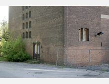 Ehemaliges Backsteingebäude mit dunkler, verwitterter Tür und kleinen, vertikalen Fensteröffnungen. Im Vordergrund ein unbefestigter Platz mit einem Bauzaun und seitlich überwuchertem Grün.