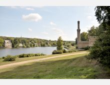 Landschaft mit einem See, umgeben von Wäldern und Wiesen. Rechts steht ein historisches Gebäude mit einem hohen Turm, während links am Ufer weitere kleinere Gebäude zu sehen sind.