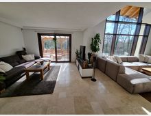 Wohnzimmer mit großen Fenstern, die den Blick auf eine bewaldete Außenlandschaft bieten. Der Raum ist mit zwei Sofas, einem Fernseher auf einem niedrigen Tisch, einem Teppich und Pflanzen dekoriert; durch eine Glastür ist eine Terrasse mit Holzüberdachung sichtbar.