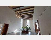 Heller Innenraum mit hoher Decke, farbigen Holzbalken, zwei großen Kronleuchtern und hohen Fenstern. In der Mitte stehen Topfpflanzen auf einem Holztisch, rechts ist eine geöffnete Tür zu sehen.