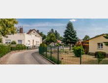 Wohnstraße in einer Vorstadt mit einer Kurve, gesäumt von Einfamilienhäusern mit roten und gelblichen Fassaden und gepflegten Gärten, die durch Metallzäune abgegrenzt sind. Im Hintergrund stehen Bäume und einige kleinere Gebäude, bei klarem Himmel.