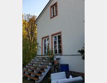 Zweigeschossiges Haus mit weißer Fassade und rötlich umrahmten Fenstern, einer Holztreppe, die zu einer Terrasse führt, sowie Pflanzen und Möbeln im Außenbereich. Im Hintergrund stehen Bäume mit herbstlichem Laub.