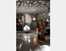 Ein moderner Innenraum mit einer Discokugel in Bodennähe, deren Lichtmuster auf Wände und Decke projiziert werden. Im Raum befinden sich dekorative Elemente wie Trockenblumen, ein großer Spiegel, gepolsterte Hocker, ein Sofa und kleine Tische.