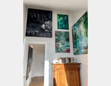 Innenraum mit einem Holzschrank im Jugendstil, darauf eine Sammlung von Büchern. An den weißen Wänden hängen mehrere großformatige, farbige Gemälde mit abstrakten Motiven, die einen hellen und modernen Kunststil zeigen. Im Hintergrund ist eine weiße Tür zu sehen.