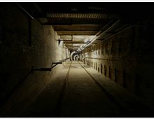 Dunkler unterirdischer Tunnel mit gemauerten Wänden, einer Schienenspur auf dem Boden und mehreren Lichtquellen an der Decke, die den Raum schwach beleuchten. Kabel und Metallstrukturen verlaufen entlang der Decke und Wände, die in die Tiefe des Tunnels führen.