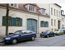 Straßenszene mit geparkten Autos vor einem historischen Wohngebäude mit cremefarbener Fassade, grünen Fensterläden und einem großen Torbogen mit blauer Holztür. Der Gehweg ist gepflastert, und ein kleiner Baum steht am Straßenrand.