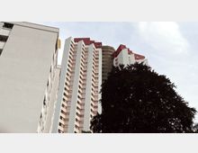 Hochhauskomplex mit mehreren weißen Gebäuden mit roten Fensterrahmen und einem zentralen runden Turm im Hintergrund, zudem ein großer Baum im Vordergrund unter einem bewölkten Himmel.